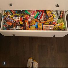 an open drawer filled with candy and other snacks on top of a wooden floor next to a person's feet