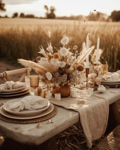 a table set with plates, napkins and flowers in front of a wheat field