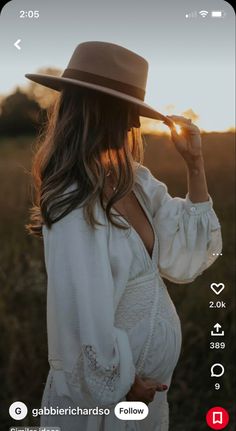 a pregnant woman wearing a white dress and hat standing in a field with the sun setting behind her