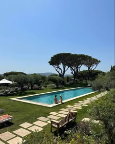 two people sitting on a bench in front of a swimming pool with trees around it