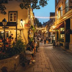 people are sitting at tables in an alleyway with lights strung over the buildings on either side