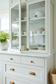 a white china cabinet with glass doors and gold handles in a kitchen, next to a wicker basket on the floor