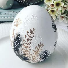 an egg decorated with flowers and leaves on a white plate next to a flower arrangement