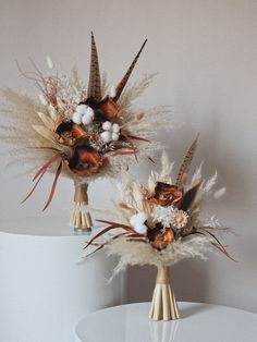 two vases with dried flowers and feathers on top of a white table in front of a wall