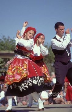 two people dressed in folk clothing dancing on the street with other people watching from behind