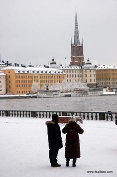 two people are standing in the snow with an umbrella over their heads and buildings behind them