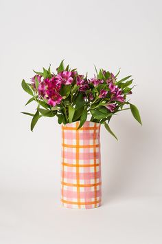 a pink and orange vase with purple flowers in it on a white surface, next to a green leafy plant