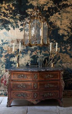 an old dresser with candles on it in front of a wallpapered wall and chandelier