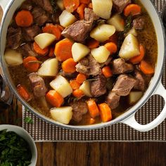 a pot filled with meat and potatoes on top of a wooden table next to a spoon