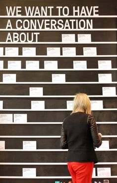 a woman standing in front of a black wall with white writing on it that says we want to have a conversation about