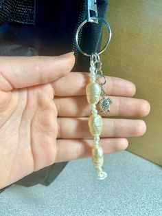 a hand holding a keychain that has shells and charms attached to it, with a bag in the background