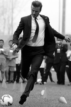 a man in a suit kicking a soccer ball on a field with other people watching