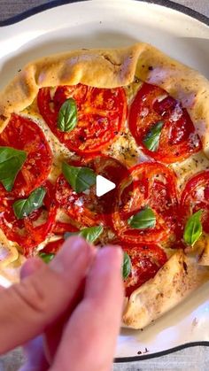 a pizza with tomatoes and basil on it in a white dish next to a person's hand