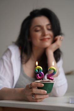 a woman sitting at a table holding a cupcake with two candles in the shape of numbers