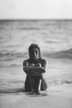 a woman is sitting on the beach with her arms around her chest and looking at the water