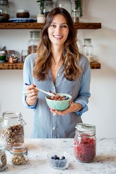 a woman holding a bowl of food in front of some jars and spoons on a table