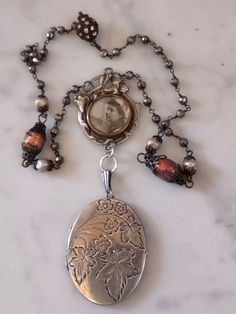 A delicate memento Mori assemblage featuring an Art Nouveau pendant from France suspending a British sterling locket with a floral chased design. The French pendant has what looks like a photo of a Normandy woman. I think the frame is silver plated, it is an antique dating around 1900. The locket is vintage. It has the interior brass bezels and plastic covers for photos. I have not taken these out to clean and there is white haze on the interior plastic. The chain is handmade pyrite rosary chain interspersed with Czech glass pearls and two antique Czech opal glass beads. Connects with a magnetic clasp. Handmade Antique Silver Pendant Locket Necklace, Vintage Etched Necklaces For Keepsake, Vintage Style Etched Necklace For Keepsake, Vintage Style Etched Necklaces For Keepsakes, Handmade Vintage Silver Locket Necklace, Handmade Brass Locket Necklace In Silver, Handmade Silver Brass Locket Necklace, Handmade Unique Silver Locket Necklace, Unique Handmade Silver Locket Necklace