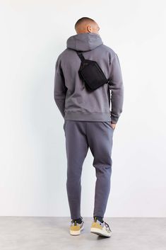 a man with a back pack standing in front of a white wall wearing grey sweatpants and a gray hoodie