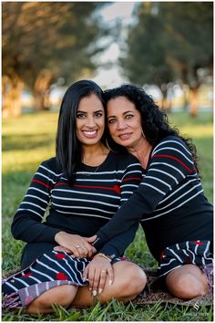 two women are sitting on the ground in front of trees and grass, one is holding her arm around the other's shoulder