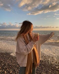 a woman standing on top of a beach next to the ocean holding a coffee cup