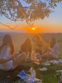 three people sitting on the grass at sunset with bottles and food in front of them