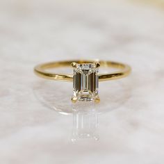 an emerald - cut diamond ring sits on a white marble surface, with its reflection in the foreground