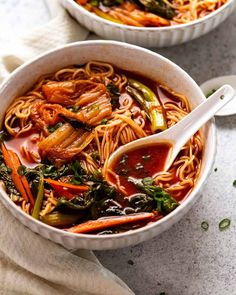 two bowls filled with noodles and vegetables on top of a white tablecloth next to spoons