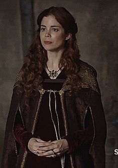 a woman with long red hair wearing a black dress and brown cape standing in front of a wall