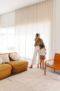 a woman standing next to a child in front of a living room with white curtains