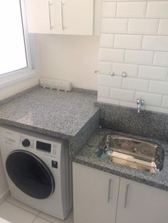 a washer sitting next to a window in a kitchen with white cabinets and gray counter tops