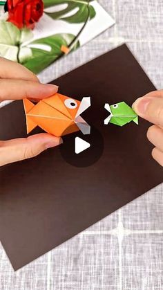 two hands holding origami fish on top of a piece of paper with flowers in the background