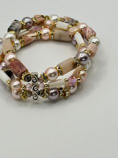 This bracelet is a multi-strand bracelet with a rich assembly of beads. The beads vary in color, shape, and texture, including glossy pink pearls and variously shaped stones with muted earthy tones. Accents of gold-tone spacers with embedded rhinestones add a touch of sparkle to the arrangement. Centerpieced with a decorative silver bar with a scroll design, which add an antique or vintage feel to the bracelet. This eclectic accessory could be a statement piece, reflecting a bohemian or artisanal style that could complement an array of outfits.   * Optional, for an extra charge you can add a set of matching earrings. The earrings  18K gold plated. A delicate matching pair of dangle earrings, each featuring a large pink pearl at the bottom. Above the pearls are a series of smaller crystals Glossier Pink, Easy Jewelry, Pink Pearls, Multi Strand Bracelet, Silver Bar, Scroll Design, Shell Jewelry, Strand Bracelet, Silver Bars