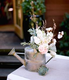 a metal watering can with flowers in it on top of a white cloth covered table