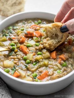 a person dipping a piece of bread into a bowl of vegetable soup with peas and carrots