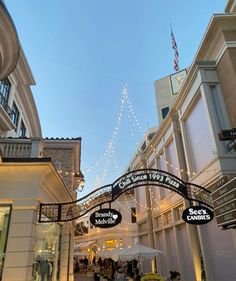 people are walking through an outdoor mall with lights strung from the ceiling and signs above them