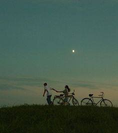 two people holding hands and walking with their bikes in the grass at dusk, while the sun is setting behind them