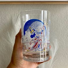 a person holding up a glass with an image of mickey mouse on the side and other items behind it