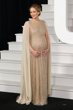 a pregnant woman wearing a gold gown and cape stands on stairs in front of a black background