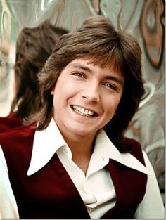 a young man smiling in front of a graffiti covered wall and wearing a maroon vest