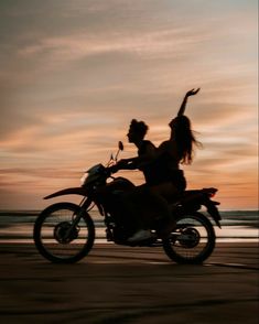 two people riding on the back of a motorcycle down a beach side road at sunset