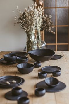 black bowls and plates on a wooden table with dried flowers in the vase behind them