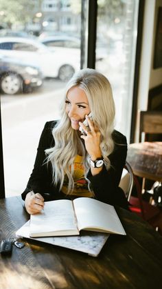 a woman sitting at a table talking on a cell phone and holding a book in her hand