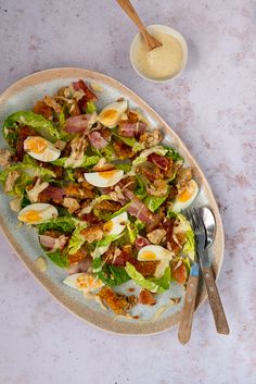 a salad with bacon and hard boiled eggs on a plate next to a bowl of dressing