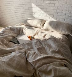 an unmade bed with a book on the bottom and white brick wall in the background