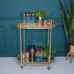 a gold bar cart with wine glasses and plants on the top, in front of a blue wall