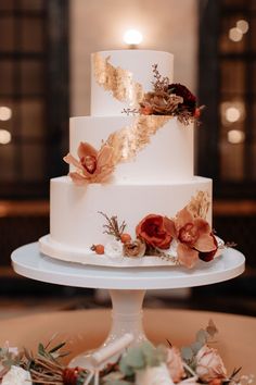 a three tiered white wedding cake with red flowers on the top and gold leaf decorations