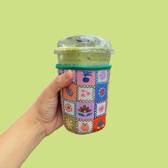 a hand holding up a cup with green liquid in it and colorful squares on the lid