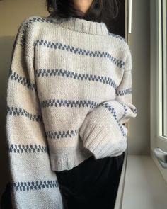 a woman wearing a white and blue sweater standing next to a window with her hands on her hips