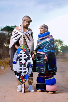 two people standing next to each other on a dirt road with one holding a surfboard