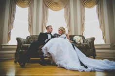 a bride and groom sitting on a couch in front of two windows with drapes